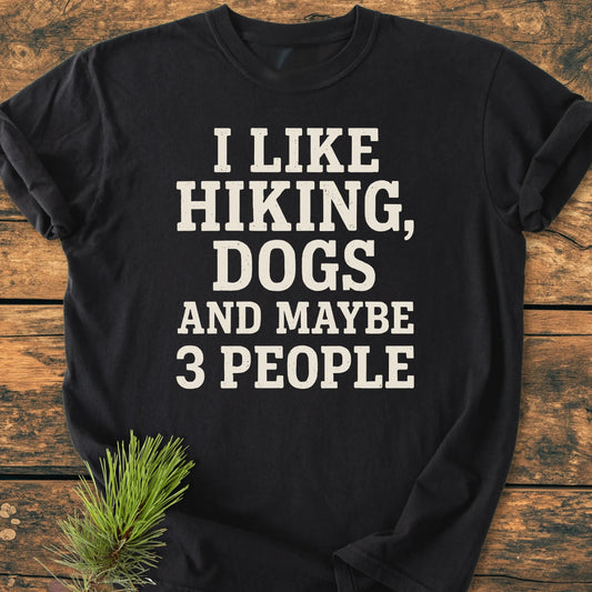 Black T-shirt, 1431 I Like Hiking And Dogs, featuring bold white text, laid flat on wood with a pine branch accent. Soft vintage wash, unisex relaxed fit, perfect for outdoor enthusiasts.