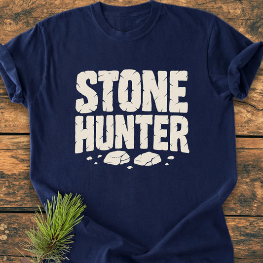 Navy-blue 1425 Stone Hunter T-shirt with cracked white lettering and stone fragments, laid flat on wood, accompanied by a pine sprig, showcasing its vintage wash and relaxed fit.