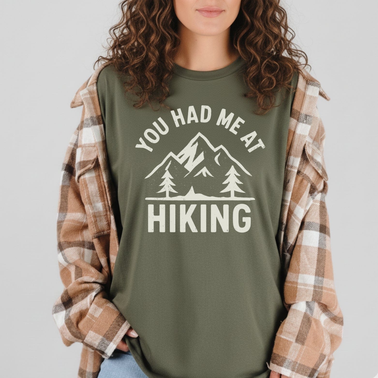 Woman wearing muted-green You Had Me At Hiking T-shirt with mountain graphic, layered under tan plaid flannel, showcasing outdoor adventure style and comfort.