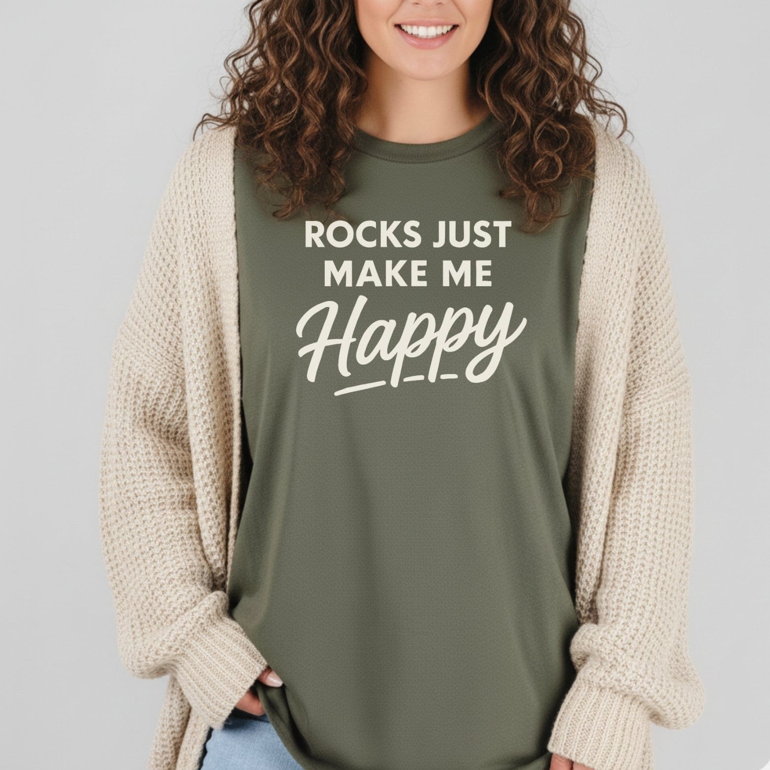 Person wearing olive-green Rocks Make Me Happy T-shirt, smiling, with loose cream cardigan, showcasing comfort and outdoor-inspired design.