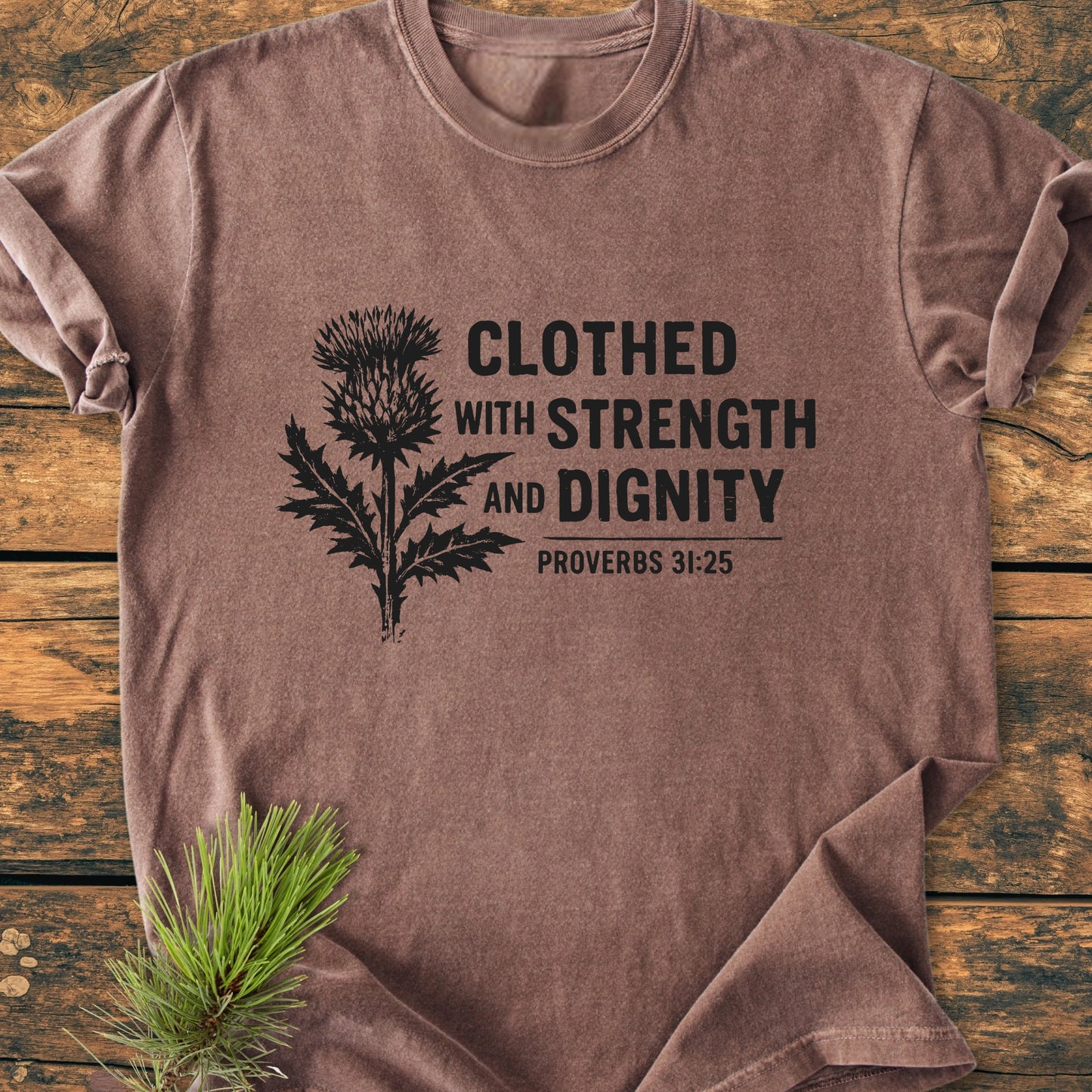 Brown short-sleeve tee laid flat, featuring Clothed With Strength And Dignity - Vintage Wash Tee with a stylized thistle and Proverbs 31:25 text, showcasing a relaxed, garment-dyed vintage finish.