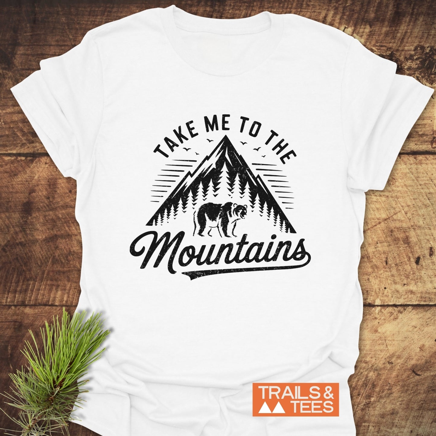 Take Me To The Mountains T-Shirt featuring a black-ink mountain design with trees, a bear, and birds, emphasizing outdoor adventure. Includes TRAILS & TEES logo.