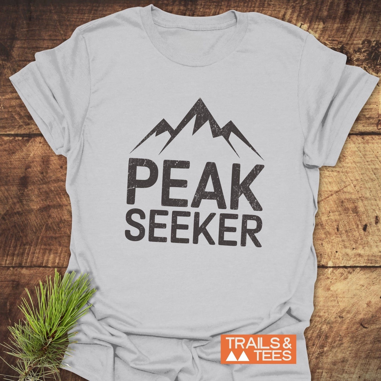 Peak Seeker T-Shirt on wood, featuring distressed lettering and mountain graphic. Includes TRAILS & TEES logo, pine sprig accent, highlighting outdoor-inspired design.
