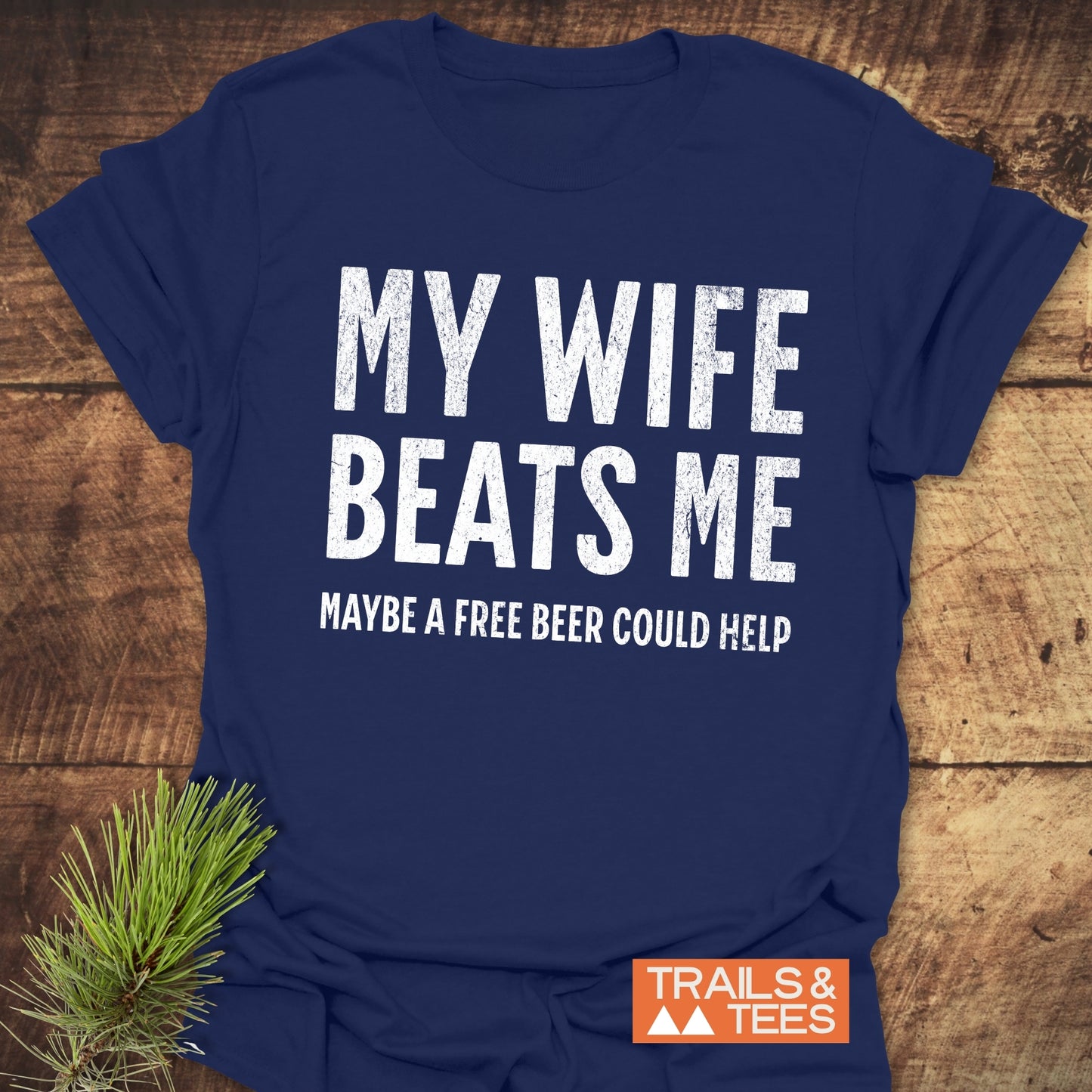 Navy-blue T-shirt with distressed white text: MY WIFE BEATS ME MAYBE A FREE BEER COULD HELP, featuring a small TRAILS & TEES logo.