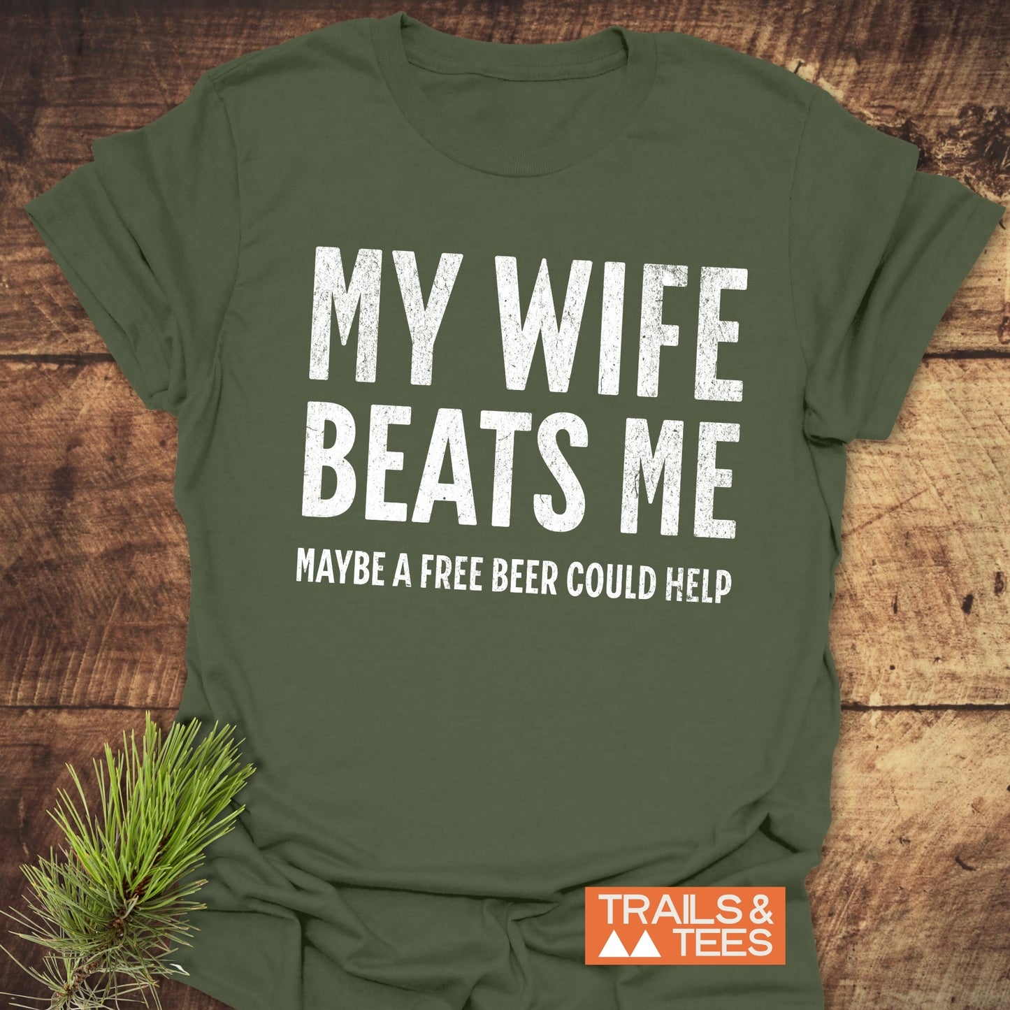 Olive-green T-shirt with My Wife Beats Me - Free Beer in distressed white text, orange TRAILS & TEES logo, pine sprig styled.