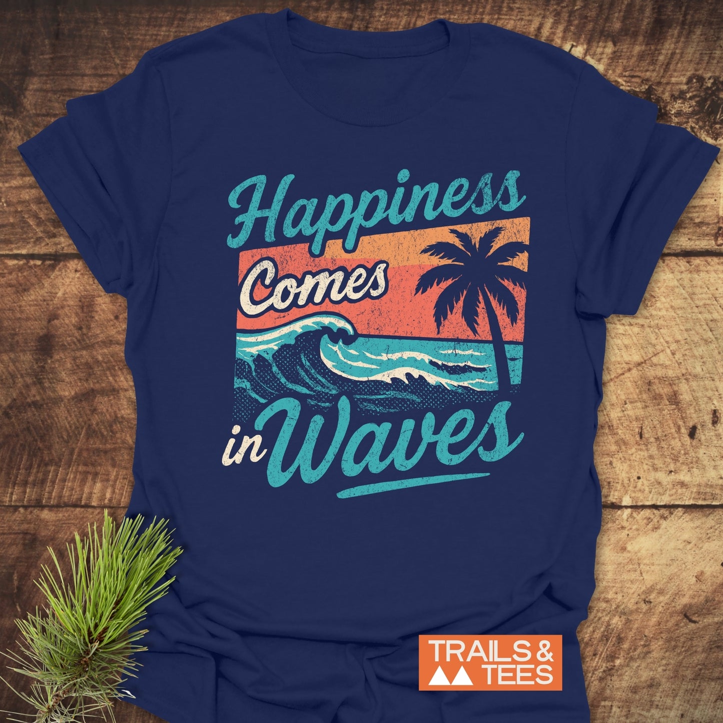 Navy-blue Happiness Comes In Waves Retro T-Shirt with sunset stripes and palm design, featuring Trails & Tees logo, laid on wood with pine sprig.