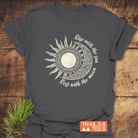 Sun & Moon Mystical Boho T-Shirt showcasing a stylized celestial motif and phrase, laid flat on wood, embodies outdoor adventure spirit with durable, comfortable cotton.