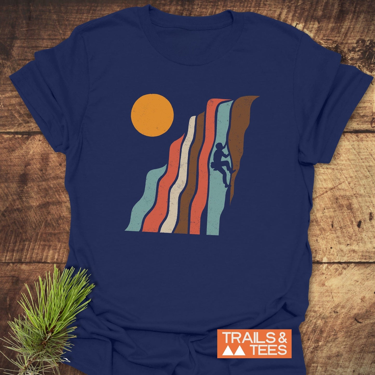 Retro Climber T-Shirt featuring a rock climber graphic with colorful vertical bands and orange sun, laid flat on wood with pine sprig.
