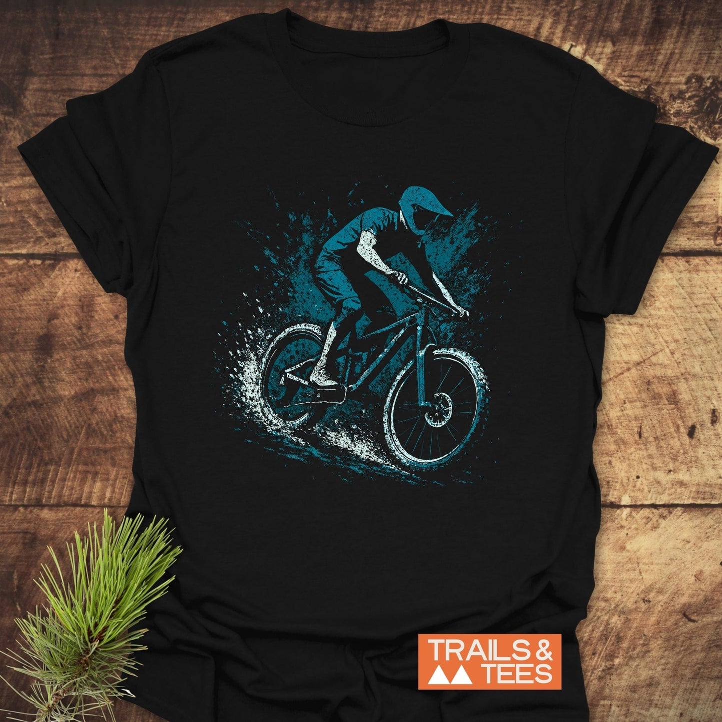 Dynamic Mountainbiker T-Shirt featuring a mountain biker graphic, crafted from soft cotton for comfort and durability, laid on a wooden surface with pine sprig.