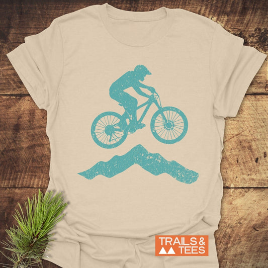 Airborne Mountainbiker T-Shirt featuring a teal mountain biker print on cream cotton, laid on wood with pine for an outdoorsy vibe.