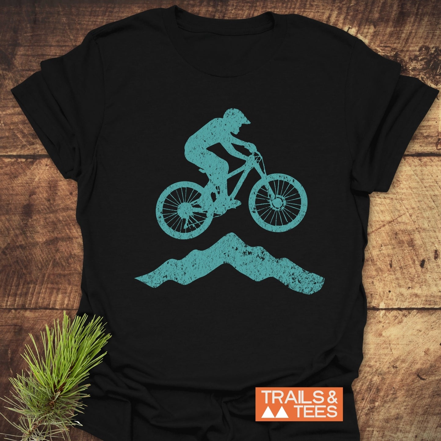 Airborne Mountainbiker T-Shirt featuring a teal biker graphic mid-jump on a black tee, set on wood, with pine sprig and TRAILS & TEES logo.