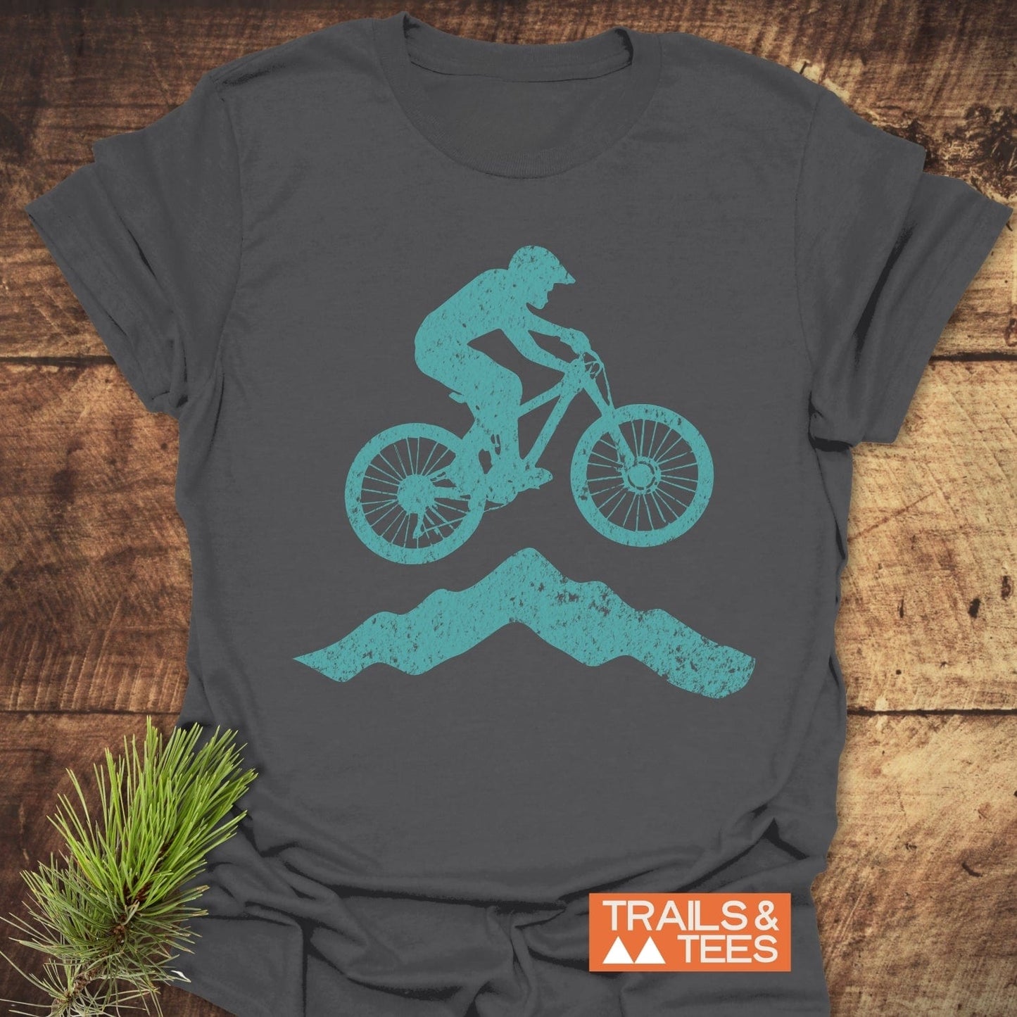 Airborne Mountainbiker T-Shirt featuring a teal biker graphic on charcoal-gray cotton, laid flat with a TRAILS & TEES logo. Perfect for outdoor adventures.