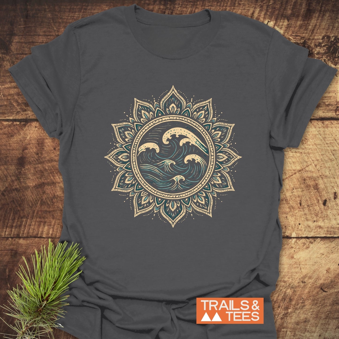 Ocean Mandala T-Shirt on wooden surface, featuring cream and teal ocean wave graphic within lotus mandala pattern. Includes “TRAILS & TEES” logo and pine sprig accent.