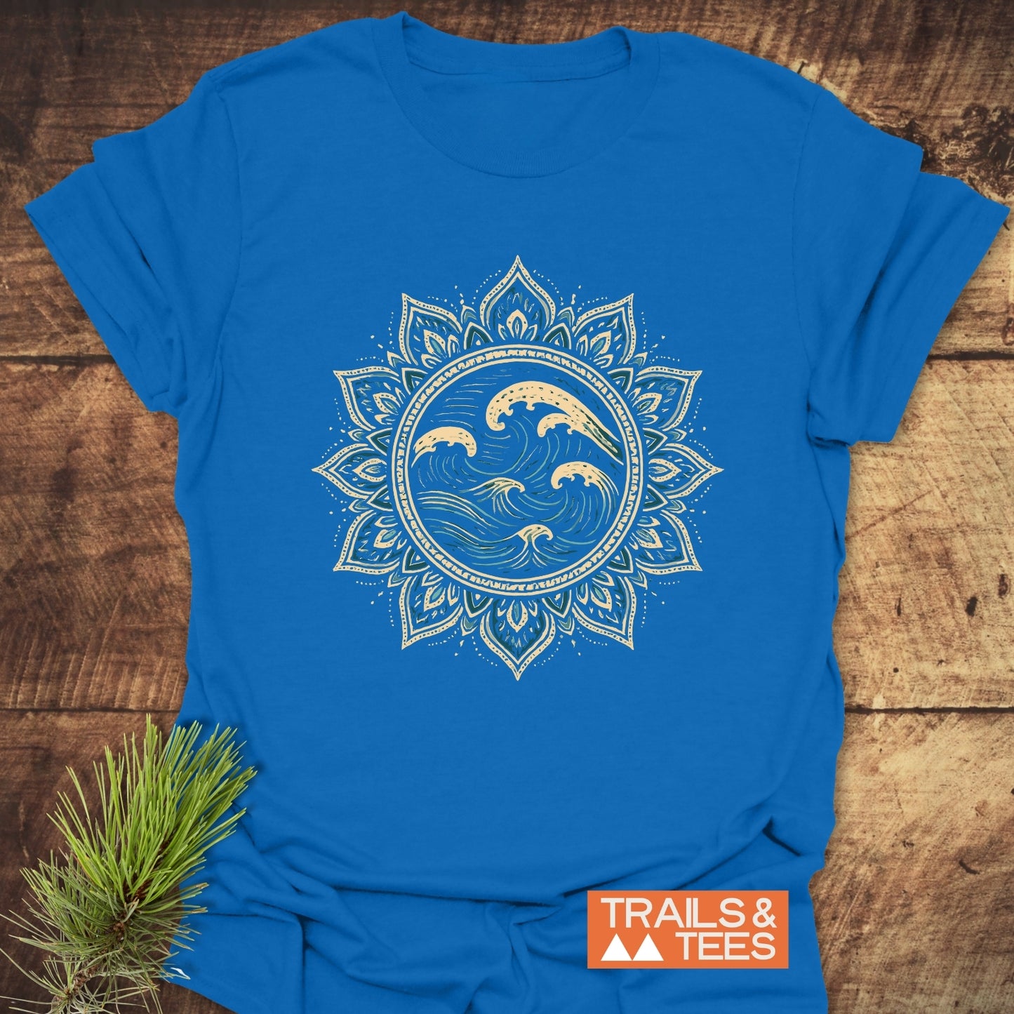 Ocean Mandala T-Shirt featuring a cream mandala graphic on bright blue, laid on wood with pine sprig, showcases outdoor-inspired design and comfort.