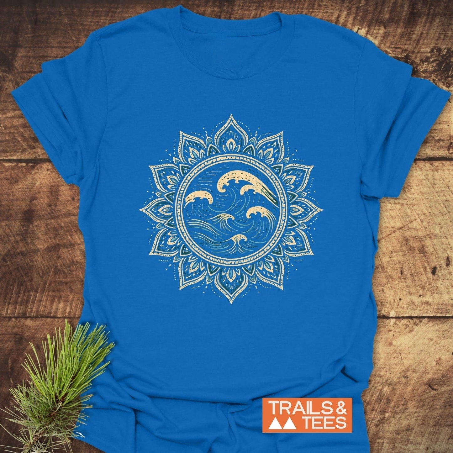 Ocean Mandala T-Shirt featuring a cream mandala graphic on bright blue, laid on wood with pine sprig, showcases outdoor-inspired design and comfort.