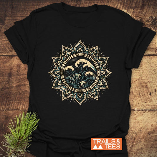 Ocean Mandala T-Shirt featuring a beige and teal mandala design with ocean waves on a black background, laid on wood, with TRAILS & TEES logo visible.