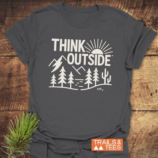 Think Outside Hiking T-Shirt featuring a white outdoor scene print, orange Trails & Tees logo, and pine sprig on a wooden surface.