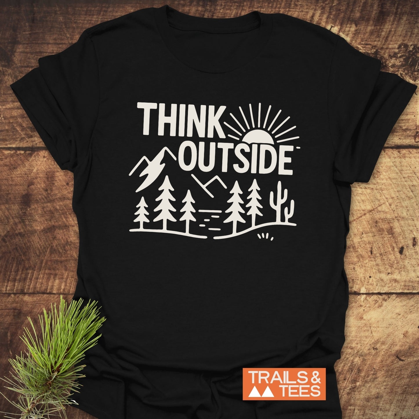 Think Outside Hiking T-Shirt features a line-art landscape with mountains and a sun, emphasizing outdoor adventure. Includes an orange TRAILS & TEES label.