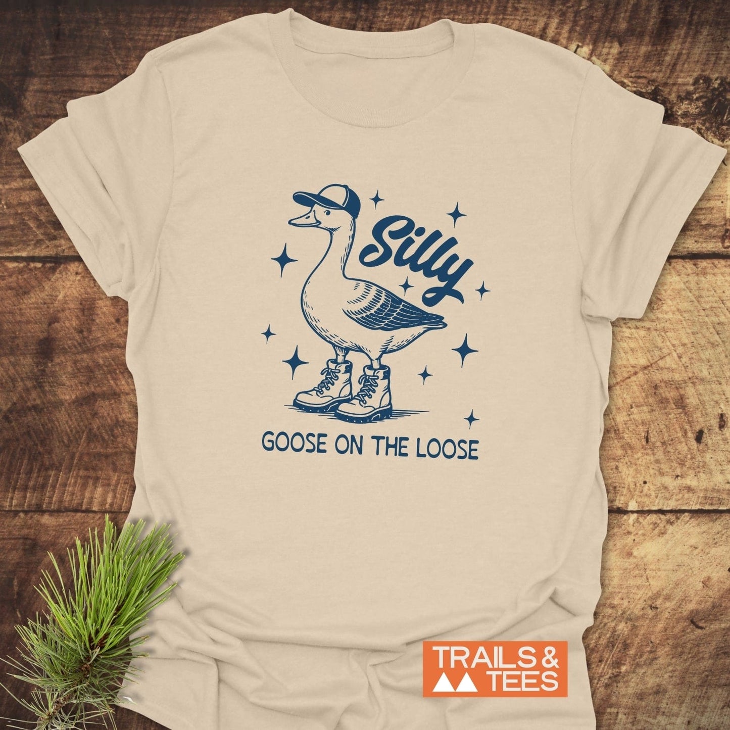 Silly Goose T-Shirt featuring a hand-drawn goose in a cap and boots, with star sparkles and “Goose on the Loose” text, laid on wood.
