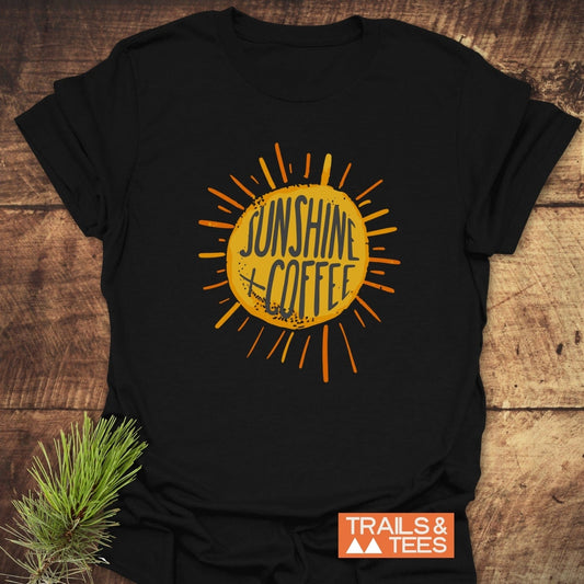 Sunshine + Coffee T-Shirt featuring a yellow-orange sunburst graphic, crafted from soft cotton, laid on wood with a pine branch accent.