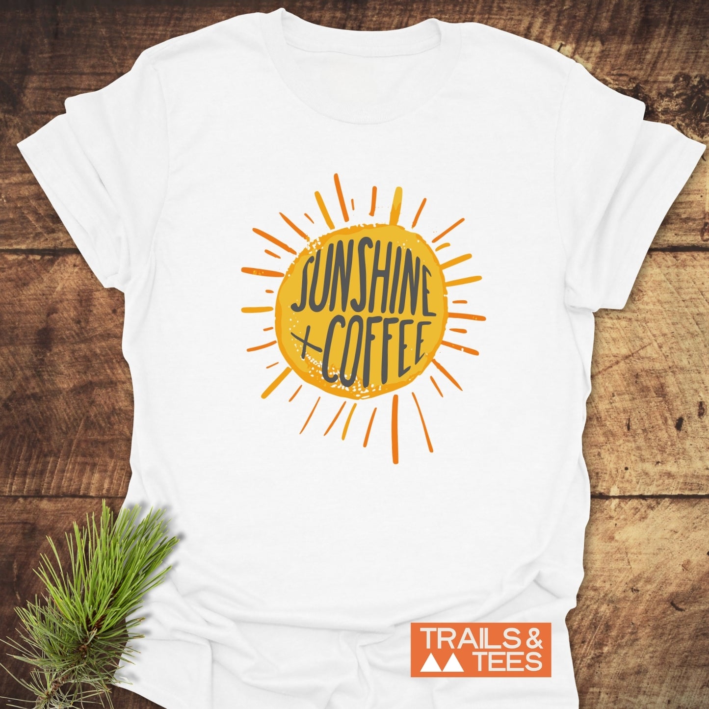 Sunshine + Coffee T-Shirt featuring a hand-drawn sunburst graphic, laid flat on a wooden surface, highlighting its soft, lightweight design for outdoor adventures.