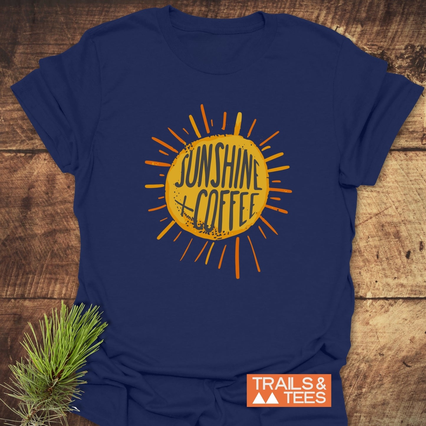 Navy-blue Sunshine + Coffee T-Shirt with sunburst graphic, laid on wood, featuring durable prints and soft, lightweight cotton for outdoor adventures.