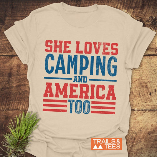 She Loves Camping T-Shirt on wood surface, features distressed red and blue slogan. Includes Trails & Tees logo, pine branch accent. Made from soft, ring-spun cotton.