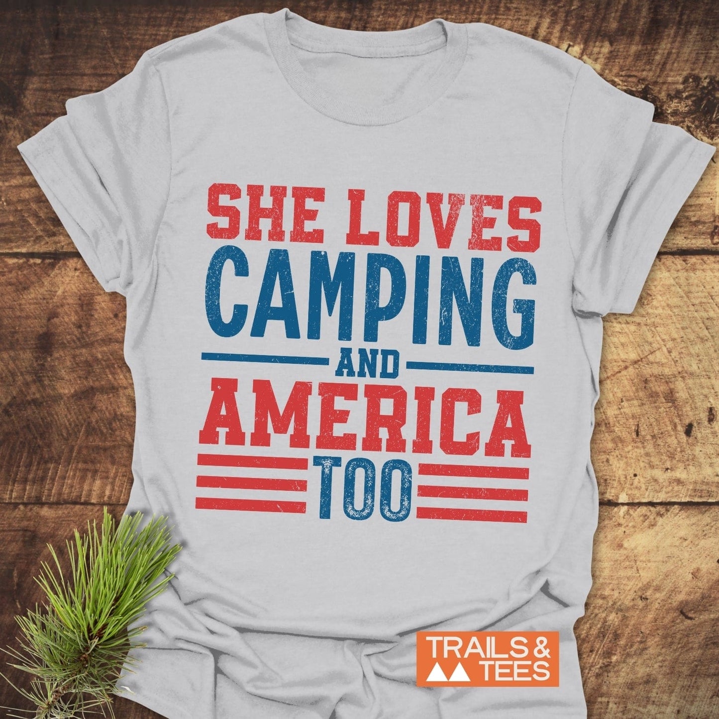 White crew-neck T-shirt with She Loves Camping and America Too slogan, laid on wood with pine branches, orange Trails & Tees logo visible.