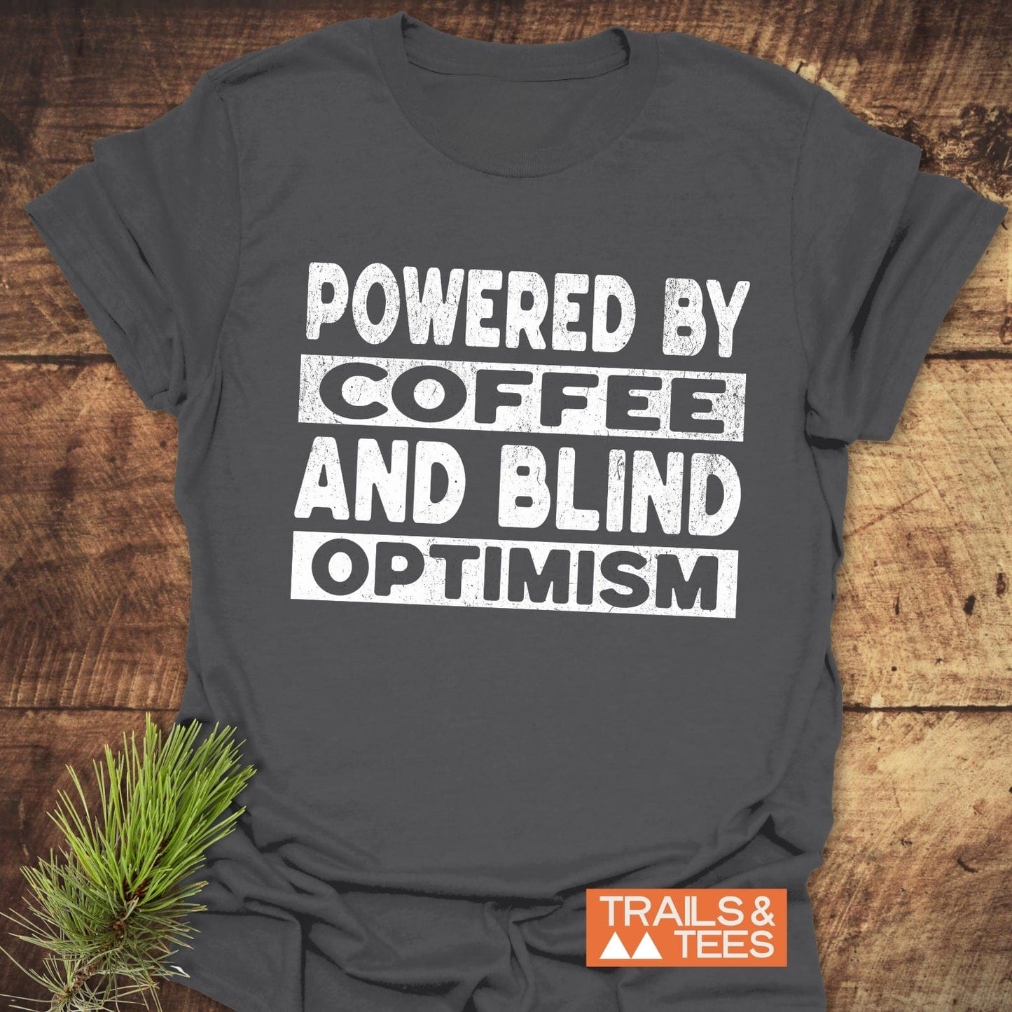 Powered By Coffee T-Shirt with white slogan on dark gray cotton, featuring a Trails & Tees logo. Laid on wood surface with pine sprig accent.