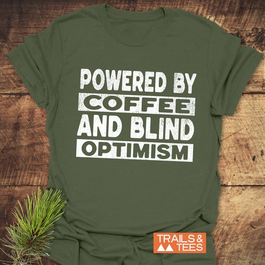 Olive-green T-shirt with POWERED BY COFFEE AND BLIND OPTIMISM slogan, featuring TRAILS & TEES logo, pine sprig, showcasing durable, adventure-ready design.