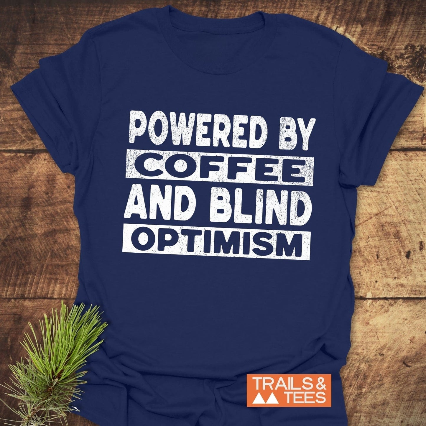 Navy-blue Powered By Coffee T-shirt with white slogan, laid on wood. Features durable print, ring-spun cotton comfort, and Trails & Tees logo.