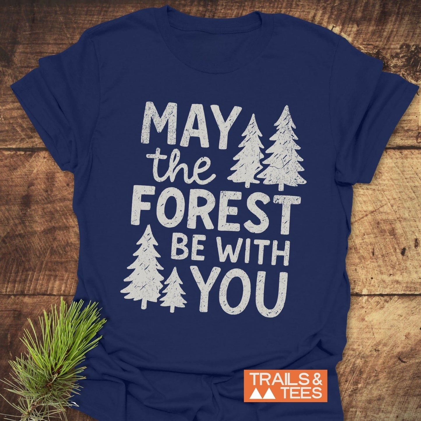Navy blue May The Forest Be With You T-shirt with white print, pine-tree graphics, and orange TRAILS & TEES logo, featuring evergreen sprig beside it.