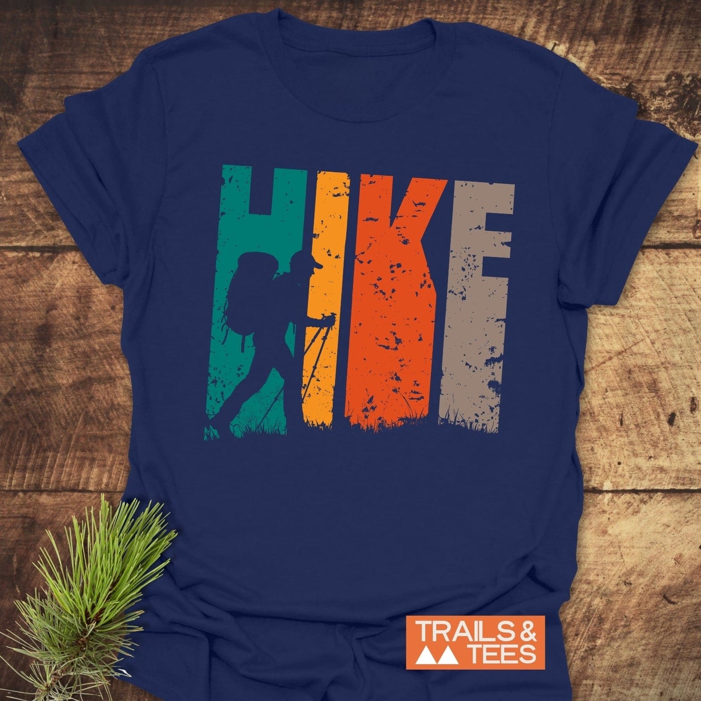HIKE Retro Sunset T-Shirt with “HIKE” in distressed blocks and a hiker silhouette, featuring a small TRAILS & TEES logo. Laid on wood with pine sprig.