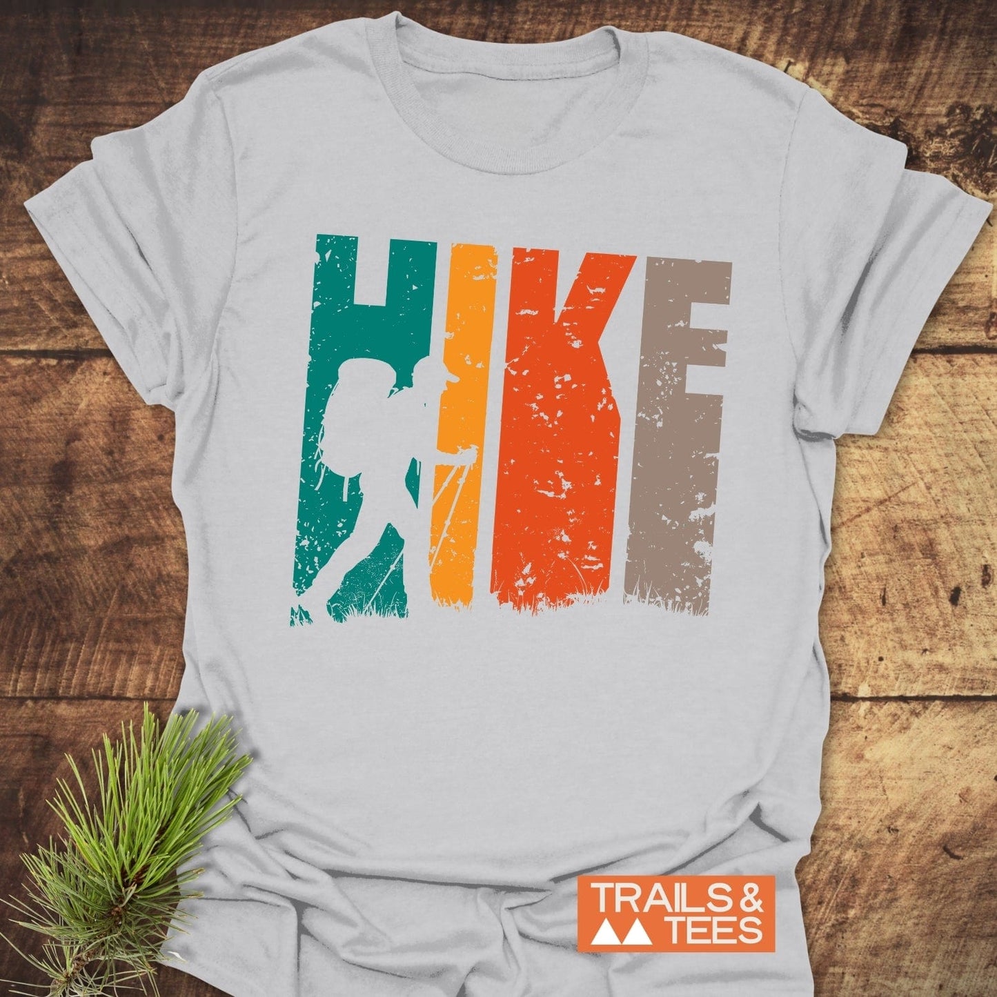 HIKE Retro Sunset T-Shirt on wood surface, featuring HIKE in earthy tones with hiker silhouette. Pine sprig and TRAILS & TEES logo visible.