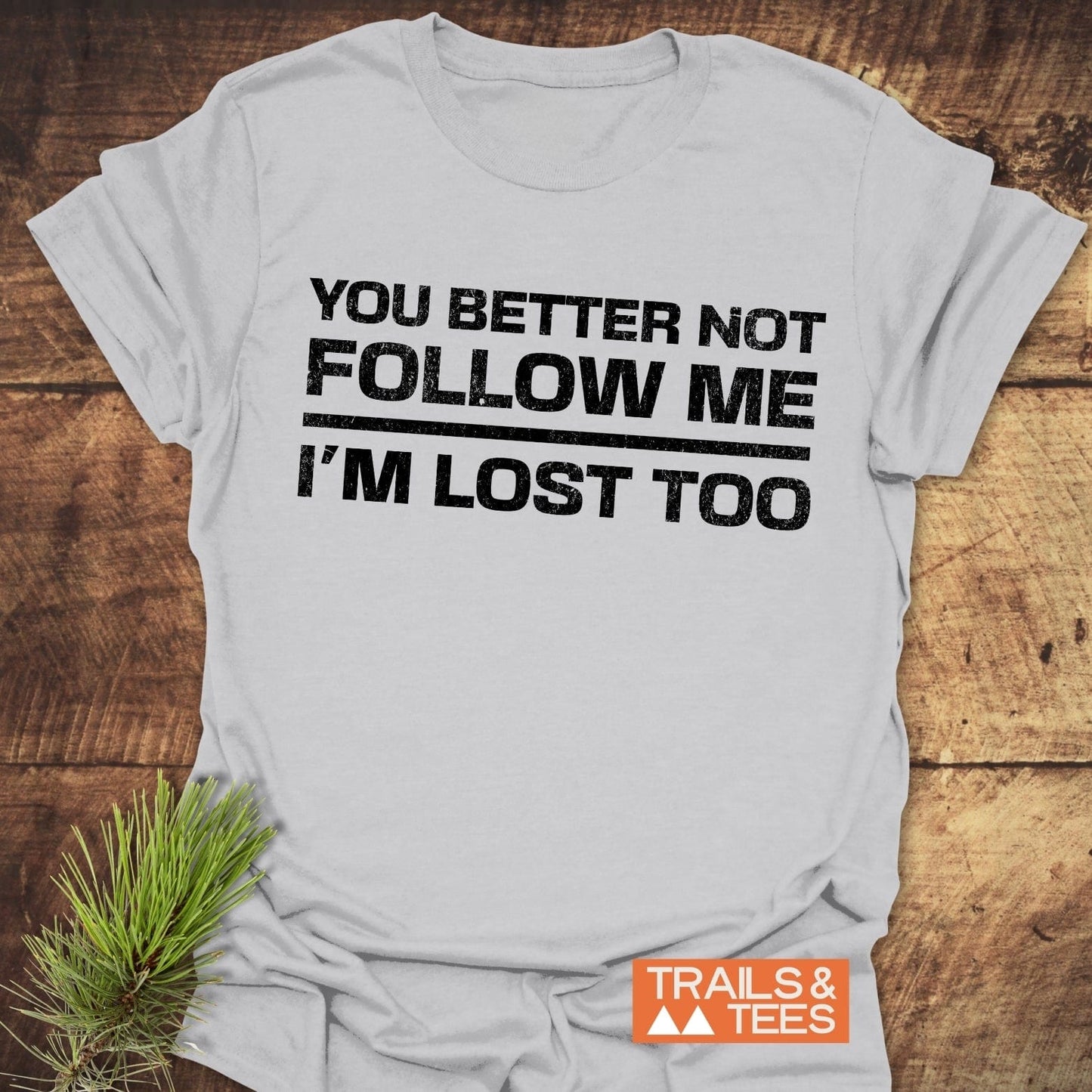 You Better Not Follow Me T-Shirt on rustic wood, featuring bold black text and Trails & Tees logo, styled with a pine needle sprig.