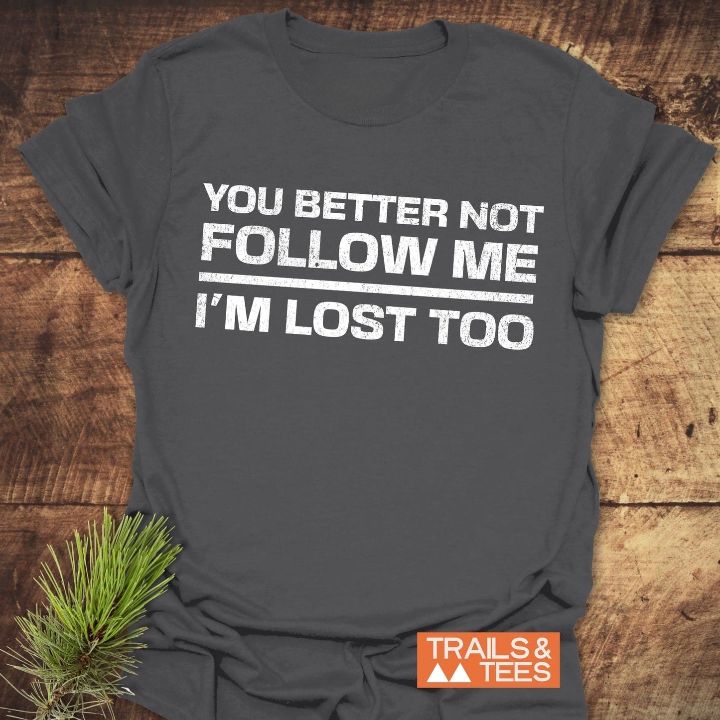 Charcoal-gray T-shirt with YOU BETTER NOT FOLLOW ME I'M LOST TOO slogan, featuring TRAILS & TEES logo, styled with a pine branch.