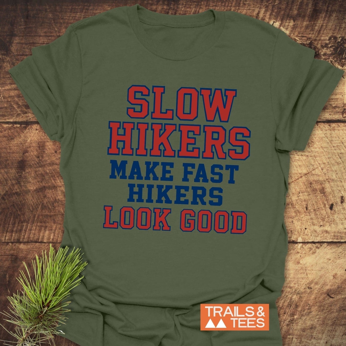 Slow Hikers Make Fast Hikers Look Good T-Shirt on wooden surface, showcasing bold text design with Trails & Tees logo and pine branch accent.