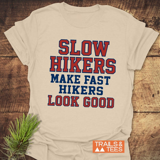Slow Hikers Make Fast Hikers Look Good T-Shirt on wooden surface, showcasing slogan. Includes Trails & Tees logo. Ideal for outdoor enthusiasts and adventure lovers.