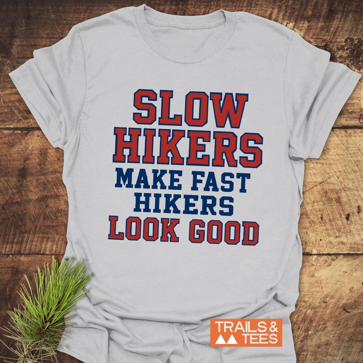 Slow Hikers Make Fast Hikers Look Good T-Shirt with collegiate lettering, laid on wood, features a Trails & Tees logo and pine sprig, highlighting outdoor theme.