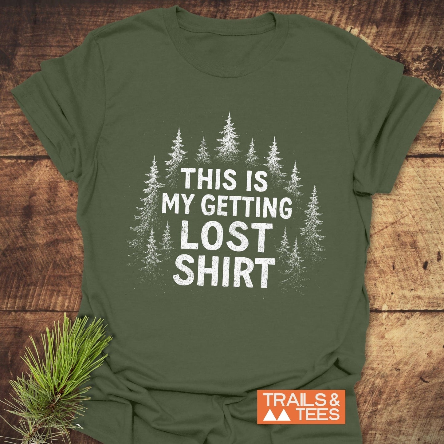 Getting Lost T-Shirt: Olive-green with white pine tree graphic and slogan, laid on wood. Includes TRAILS & TEES logo, pine sprig for outdoorsy theme.