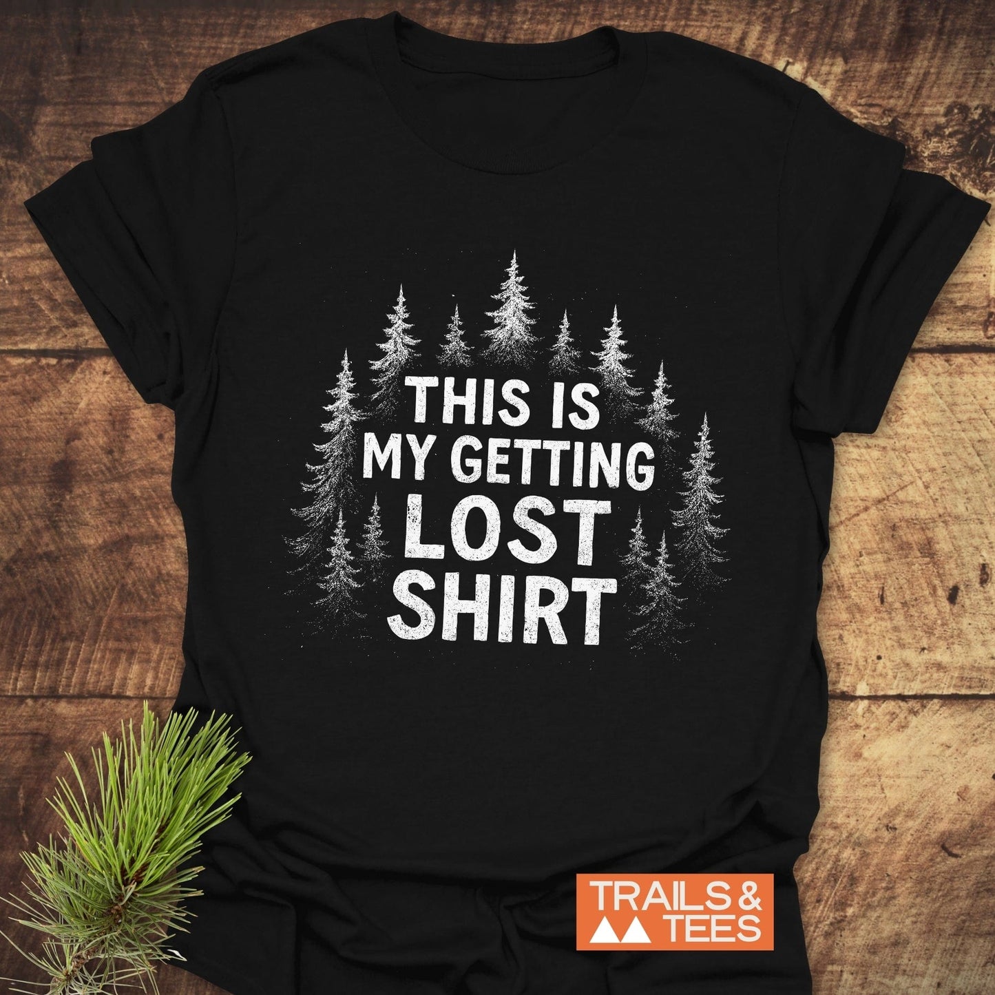 Getting Lost T-Shirt on wood, featuring evergreen trees and slogan. Includes Trails & Tees logo and a pine sprig, emphasizing an outdoor adventure theme.