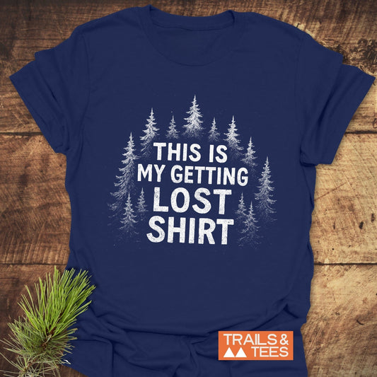 Navy-blue Getting Lost T-Shirt with white pine tree design and TRAILS & TEES logo, laid on wood, featuring soft, durable cotton suitable for outdoor adventures.
