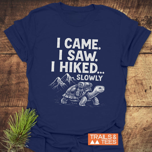 Navy blue I Came, I Saw T-Shirt featuring a tortoise design and hiking caption, displayed on wood with a pine sprig, emphasizing outdoor adventure.