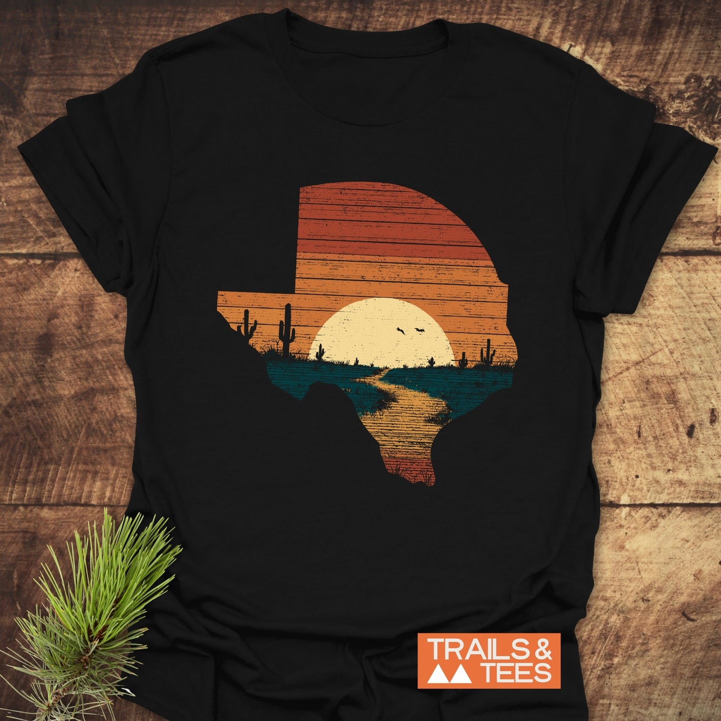 Texas Sunset T-Shirt featuring a stylized Texas silhouette with desert scene, laid on wood. Includes TRAILS & TEES logo and pine branch accent.