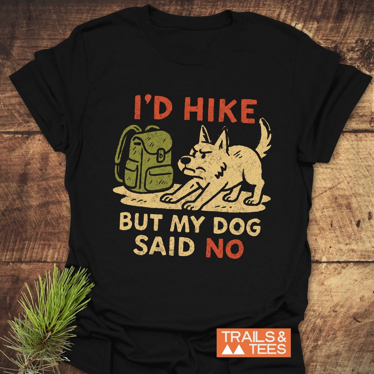 Dog Stops The Hike T-Shirt with a distressed backpack and cartoon dog graphic, slogan, and Trails & Tees logo, laid flat on a wooden surface.
