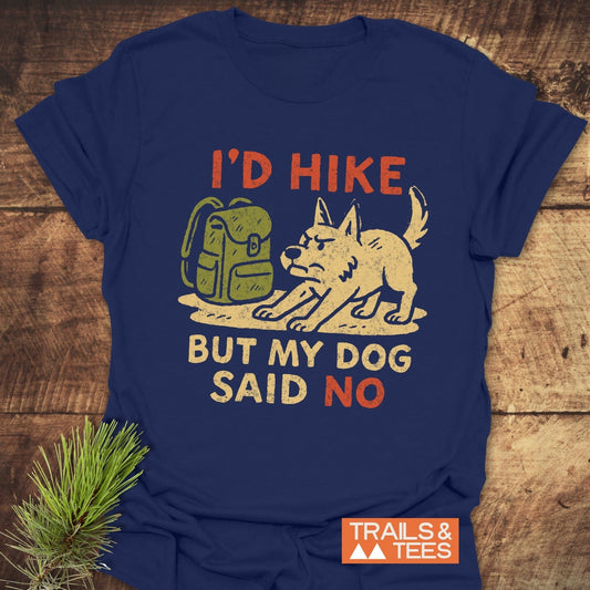 Dog Stops The Hike T-Shirt with cartoon dog and backpack design, laid on wood. Soft ring-spun cotton, ideal for outdoor enthusiasts.