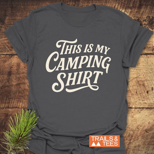 Charcoal-gray T-shirt with This Is My Camping Shirt slogan, laid on wood, featuring durable print and taped shoulders for outdoor comfort.