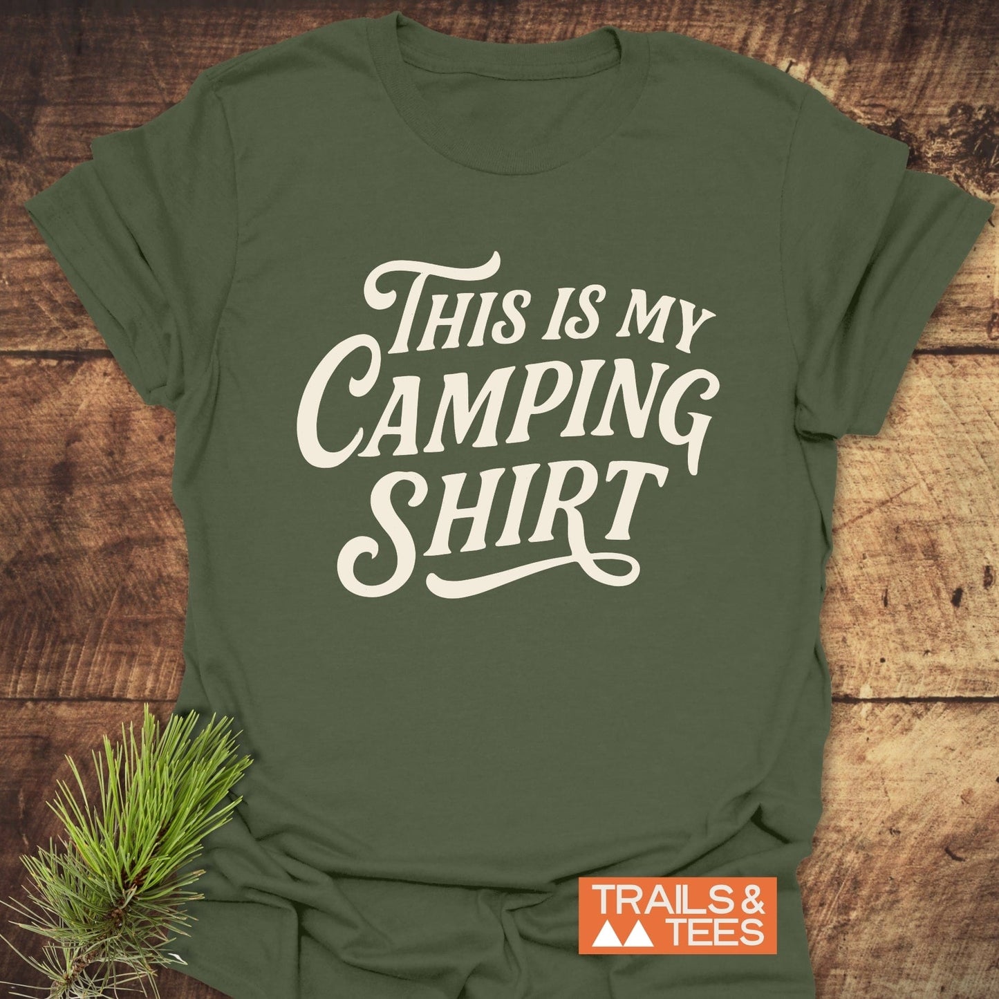 This Is My Camping T-Shirt: army-green tee with cream script, orange tag 'TRAILS & TEES,' laid on wood, featuring durable, soft ring-spun cotton for outdoor adventures.