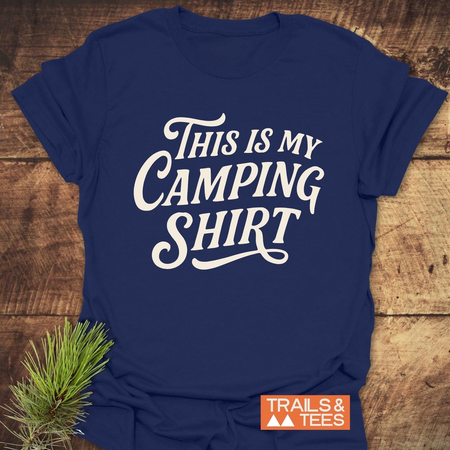 Navy-blue T-shirt with THIS IS MY CAMPING SHIRT printed in white script, featuring the TRAILS & TEES logo, laid on wooden surface with pine needles.