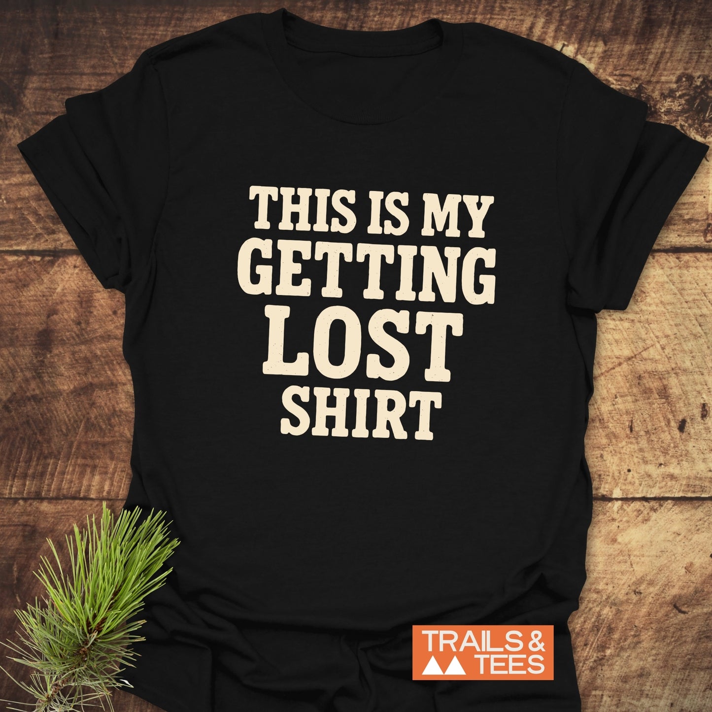 This Is My Getting Lost T-Shirt, black with off-white text, features a small orange 'TRAILS & TEES' logo, styled with a pine sprig beside it.